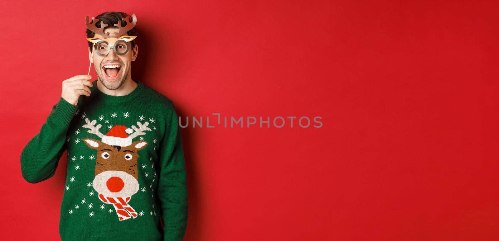 Handsome surprised man in christmas sweater, holding party mask and smiling, enjoying new year celebration, standing over red background.