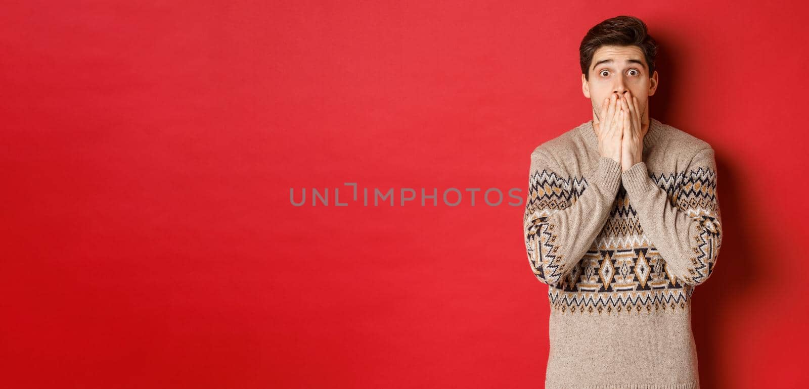 Portrait of shocked and speechless caucasian guy in christmas sweater, reacting to announcement, standing startled against red background.