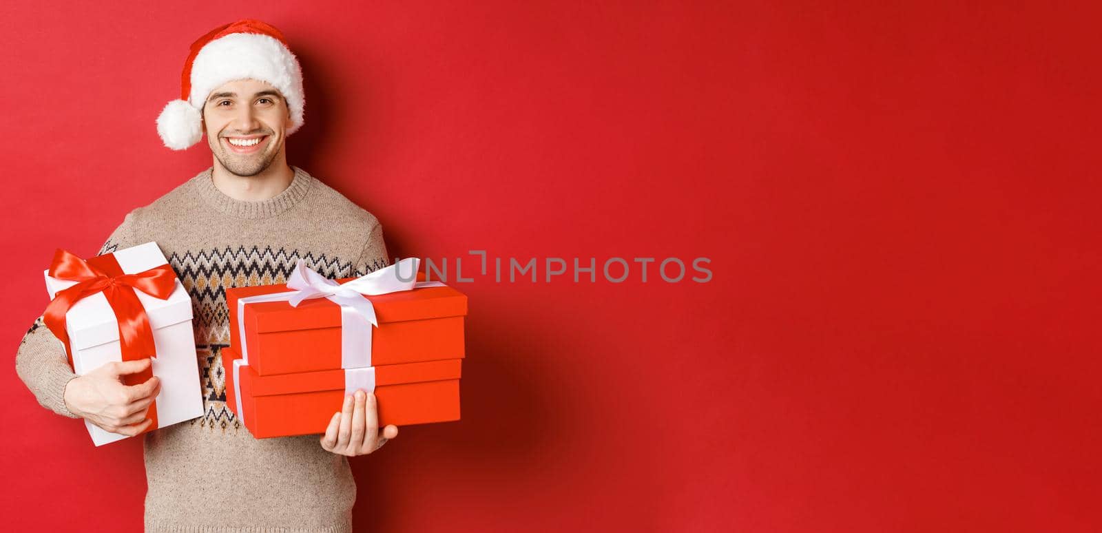 Concept of winter holidays, new year and celebration. Portrait of handsome man in santa hat and sweater, holding boxes with christmas presents and smiling, prepared gifts, red background by Benzoix