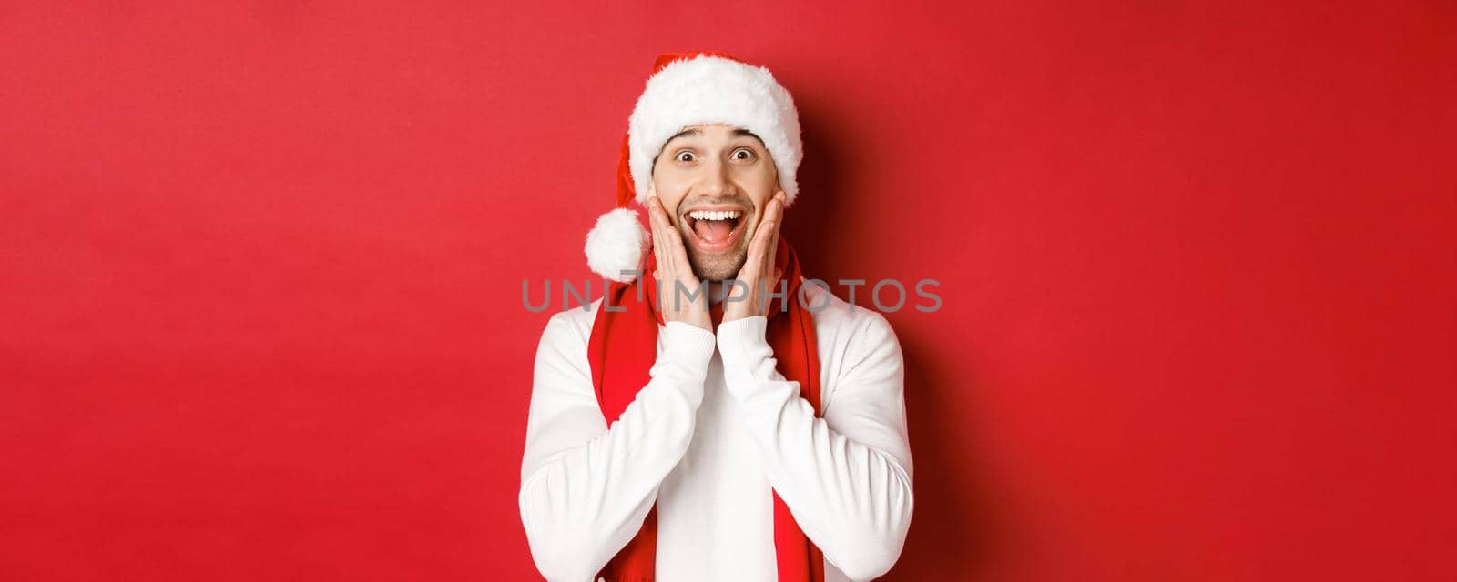 Concept of christmas, winter holidays and celebration. Close-up of surprised and happy man in santa hat and scarf, looking at something amazing, standing over red background.