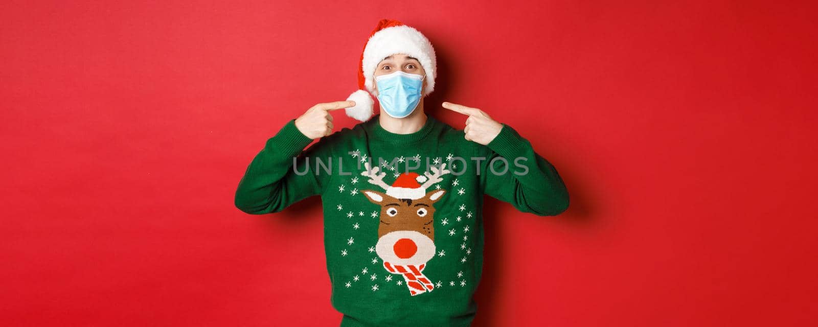 Concept of new year, coronavirus and social distancing. Happy man in santa hat and christmas sweater, recommending to wear medical mask on party, standing over red background by Benzoix