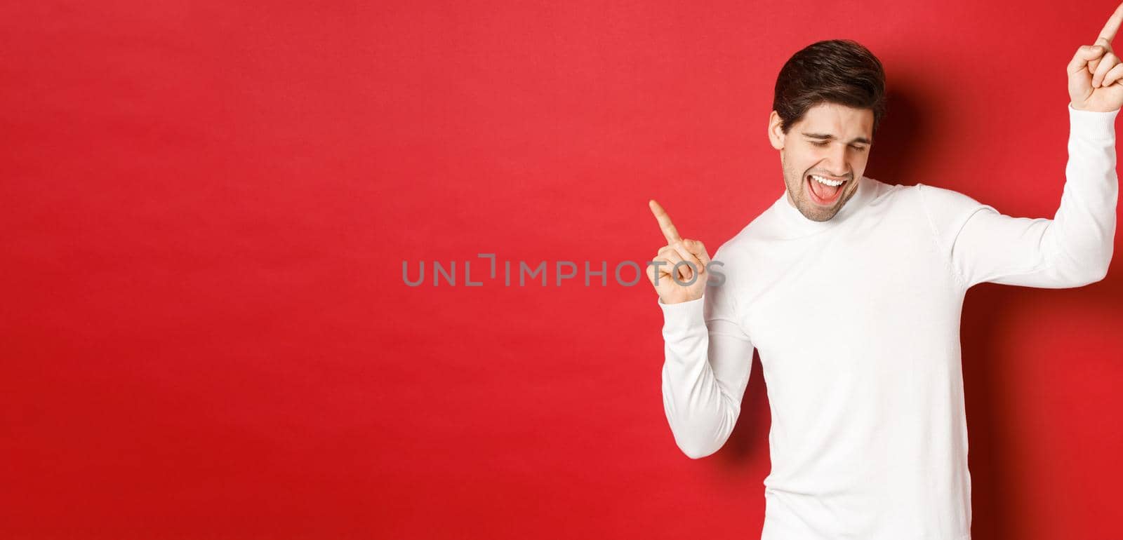 Portrait of handsome man enjoying new year party, dancing and having fun, pointing fingers up, standing over red background by Benzoix