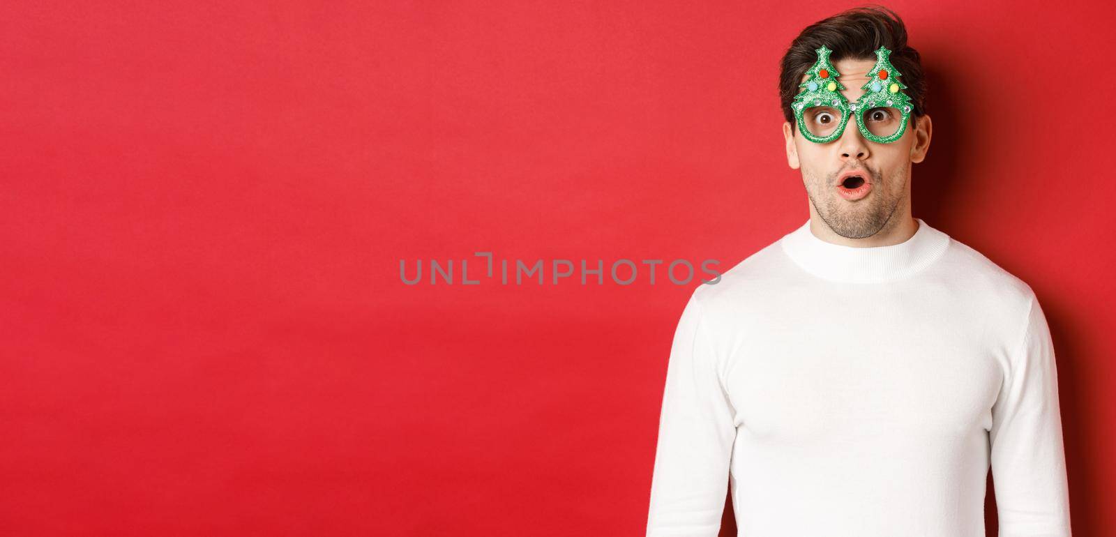 Concept of christmas, winter holidays and celebration. Close-up of surprised handsome guy in party glasses, saying wow and looking at amazing new year promo offer, red background.