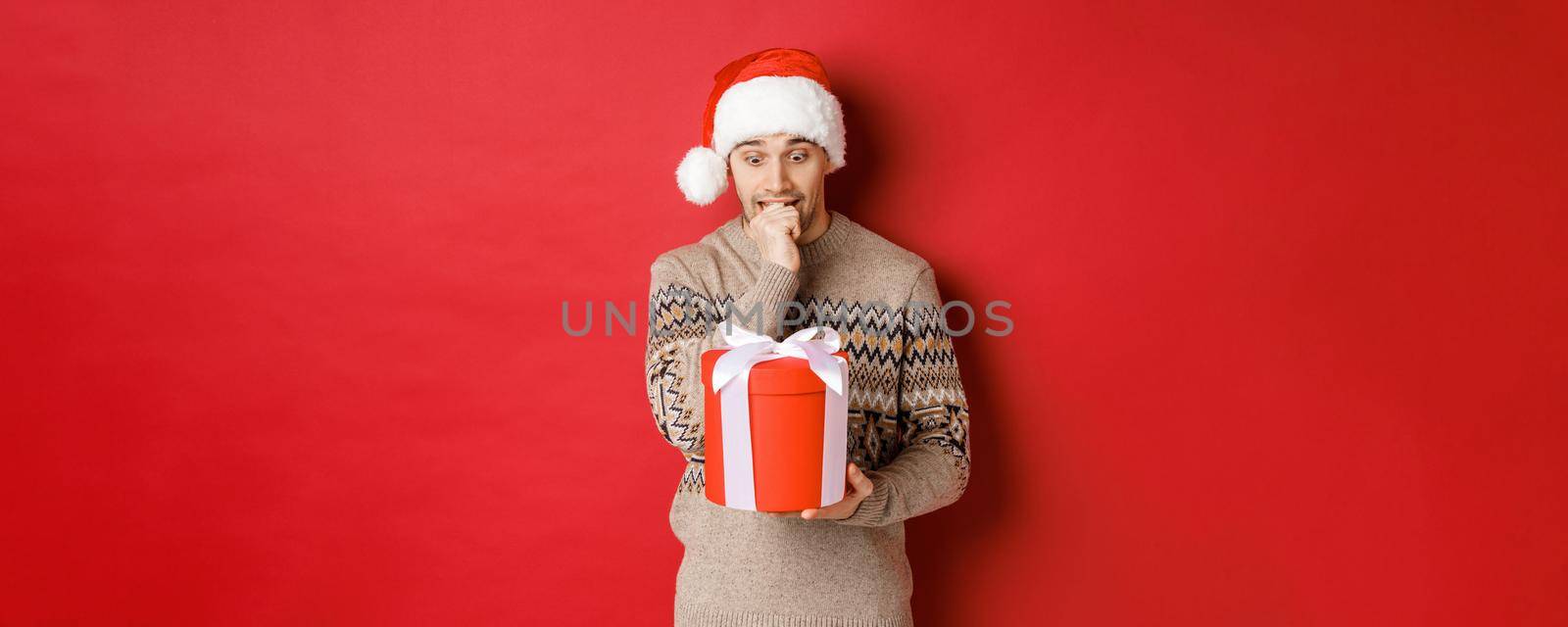 Image of worried guy in santa hat and sweater, looking indecisive at christmas gift, standing against red background.