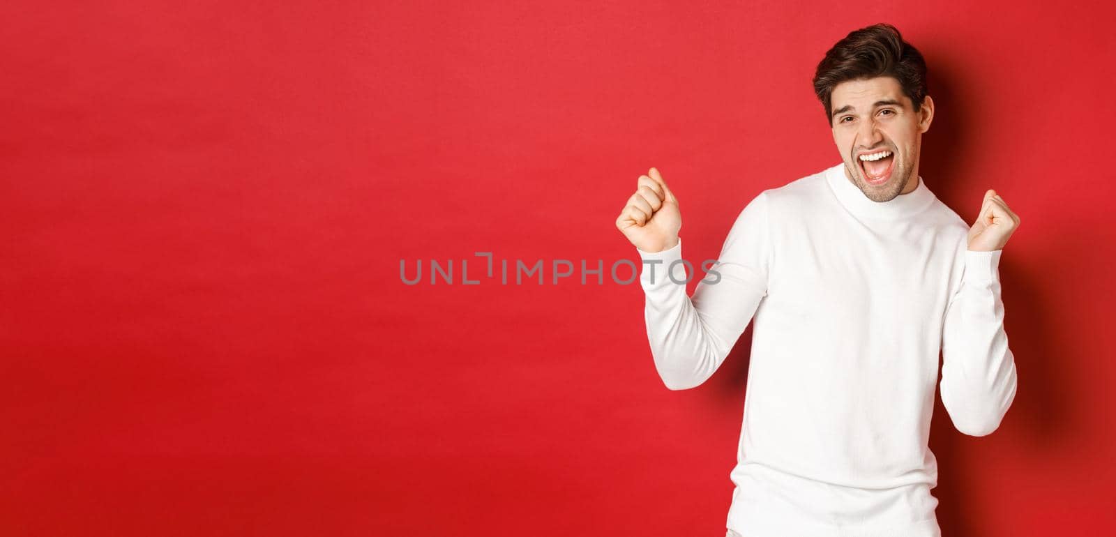Portrait of handsome man enjoying new year party, dancing and having fun, standing in white sweater against red background by Benzoix