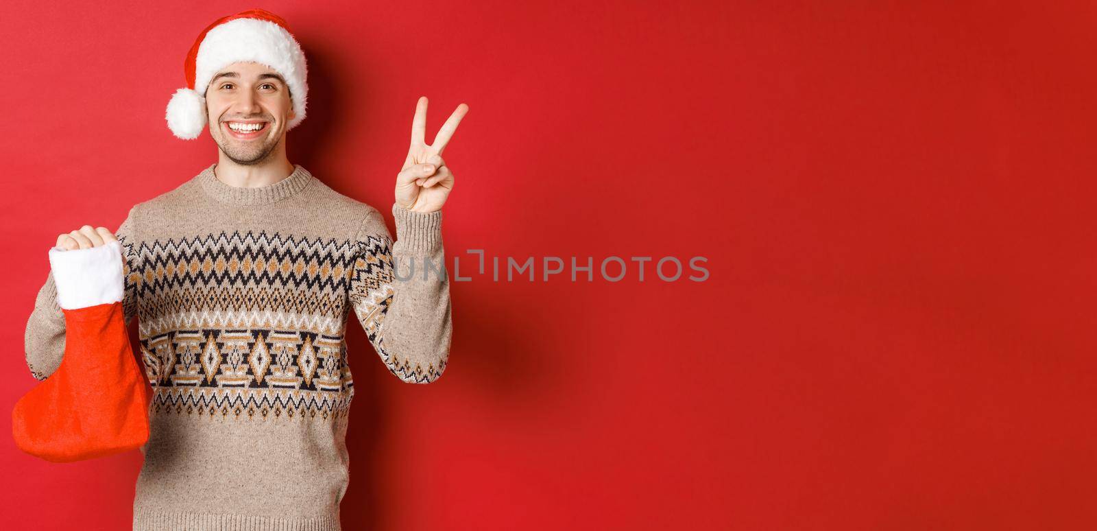 Concept of winter holidays, new year and celebration. Image of happy smiling man in santa hat and sweater, showing peace sign and a christmas stocking bag with gifts, red background by Benzoix