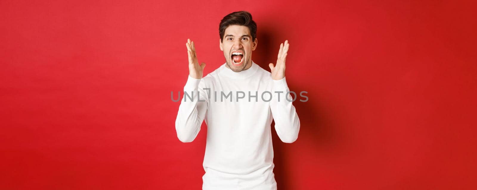 Image of frustrated and angry man in white sweater, shouting in rage, being mad at someone, standing over red background.
