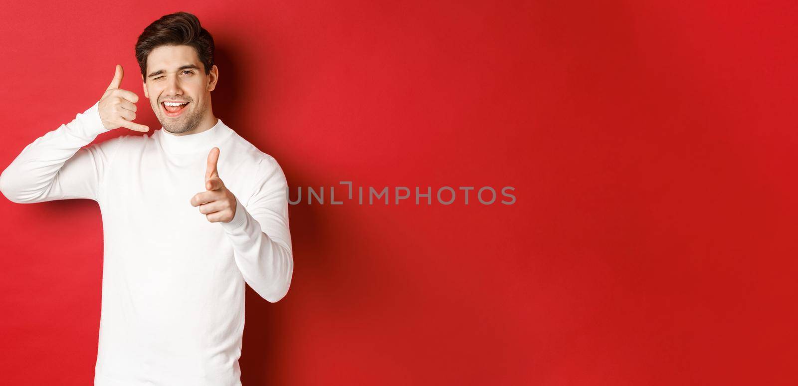 Portrait of sassy and confident handsome man, flirting with you, asking to call him, showing phone sign and pointing at camera, standing over red background.