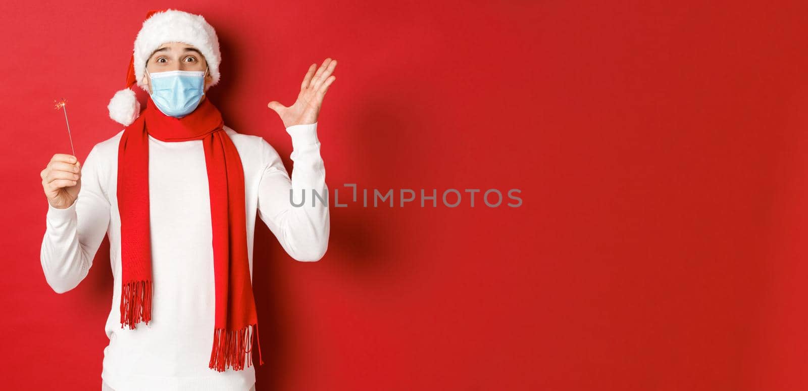 Concept of covid-19, christmas and holidays during pandemic. Cheerful handsome man in medical mask and santa hat, celebrating new year with sparkler, looking excited, red background.