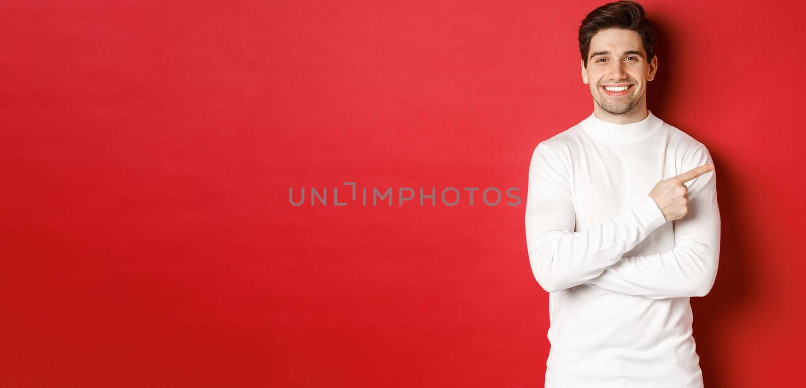 Handsome young man with bristle, wearing white sweater, pointing fingers at upper right corner and smiling, showing christmas banner, standing over red background by Benzoix