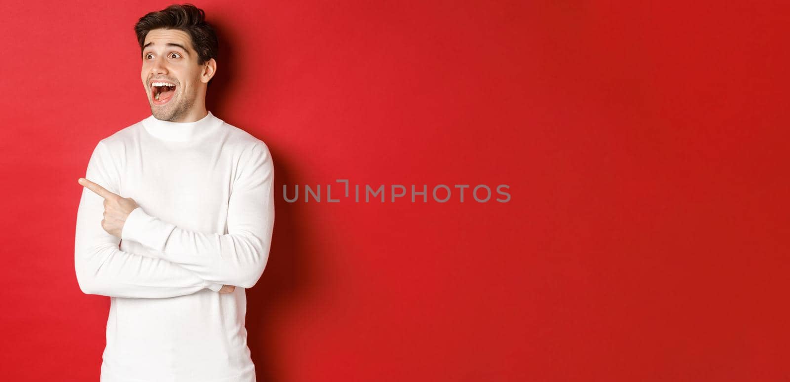 Concept of winter holidays. Image of amazed handsome man reacting to big christmas announcement, looking and pointing left excited, standing over red background.