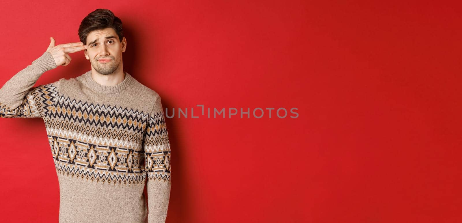 Portrait of distressed and annoyed handsome man, wearing christmas sweater, making finger gun sign near head and shooting himself, standing over red background.