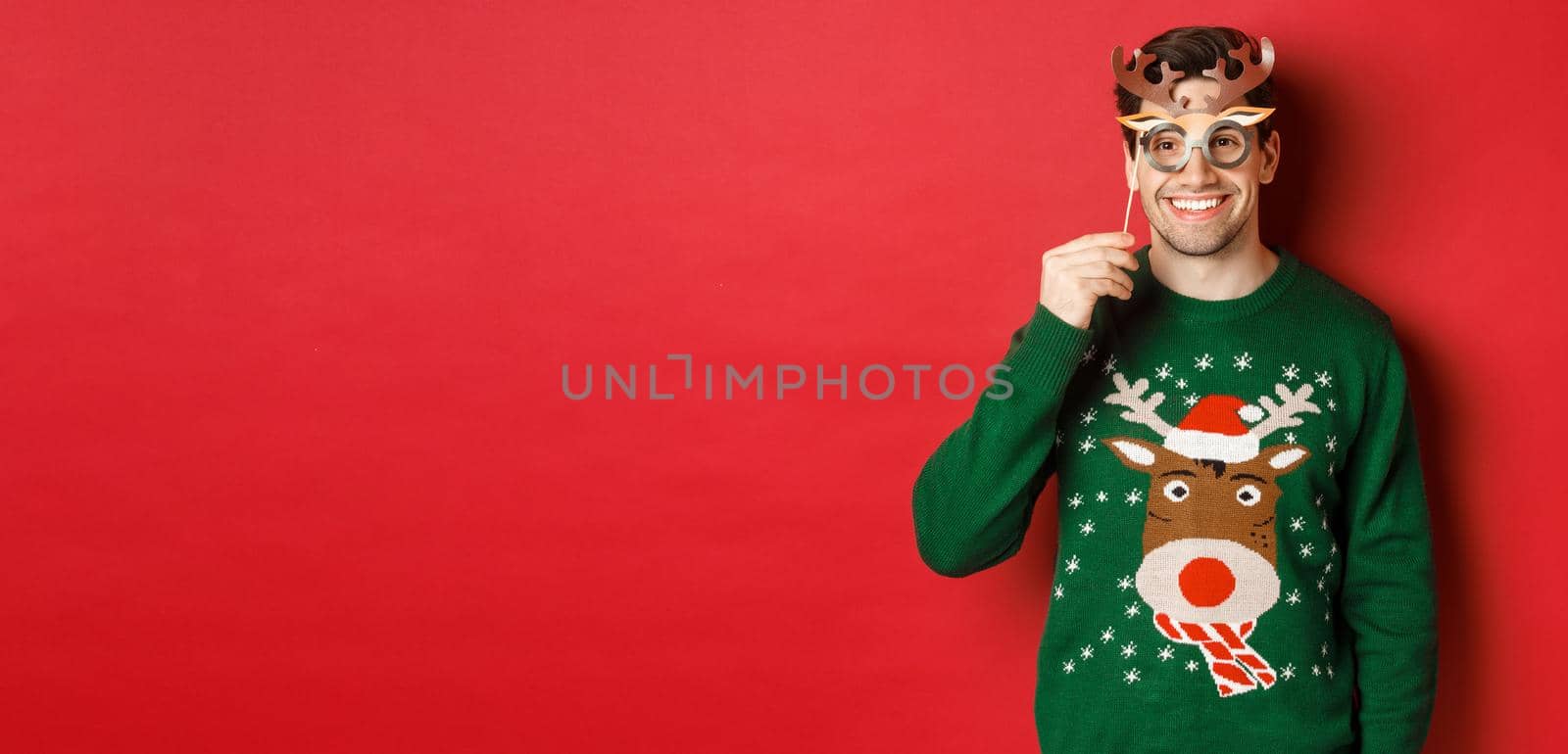 Handsome happy man in christmas sweater, holding party mask and smiling, enjoying new year celebration, standing over red background.