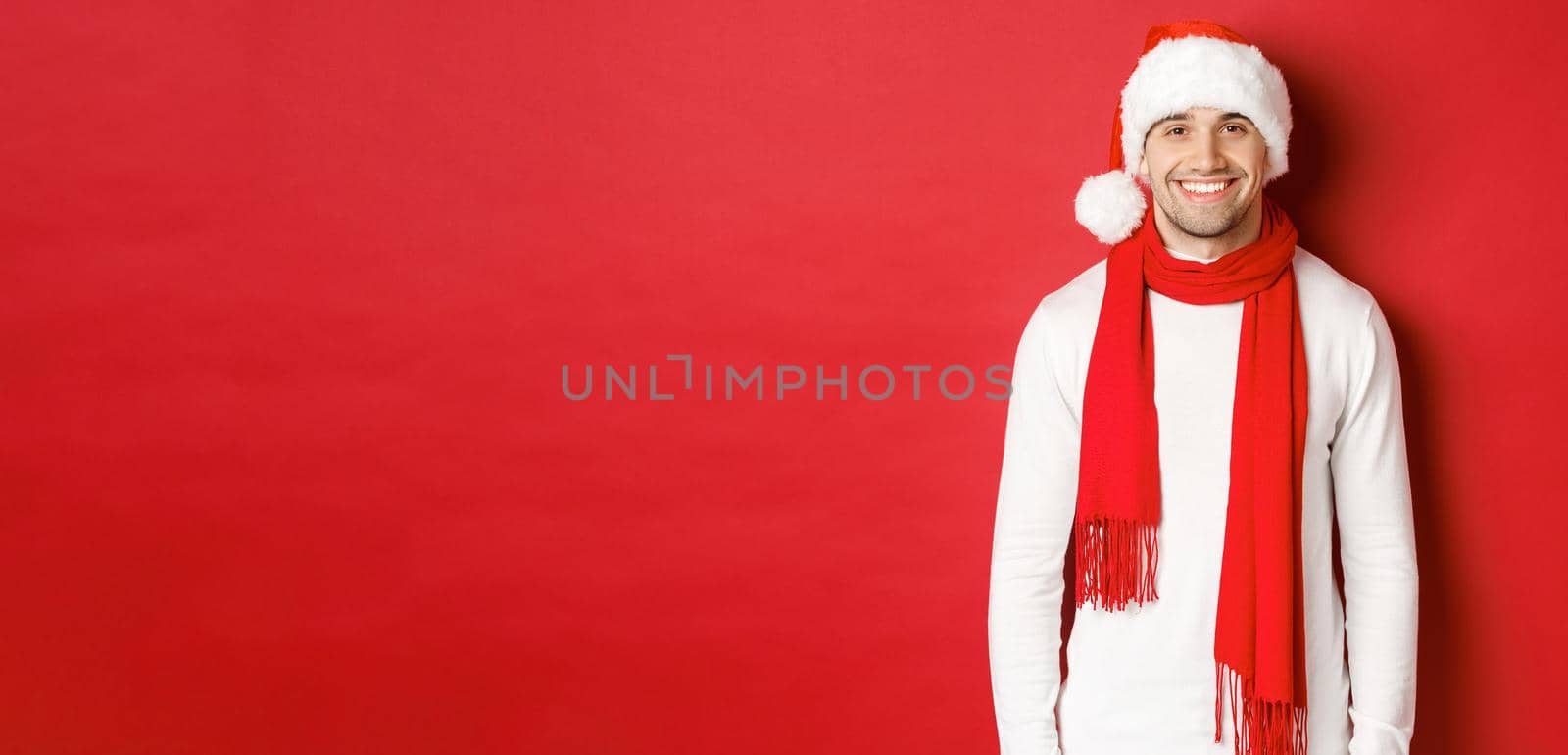 Happy caucasian guy celebrating christmas and smiling, wearing santa hat, scarf and white sweater, enjoying new year holidays, standing over red background.