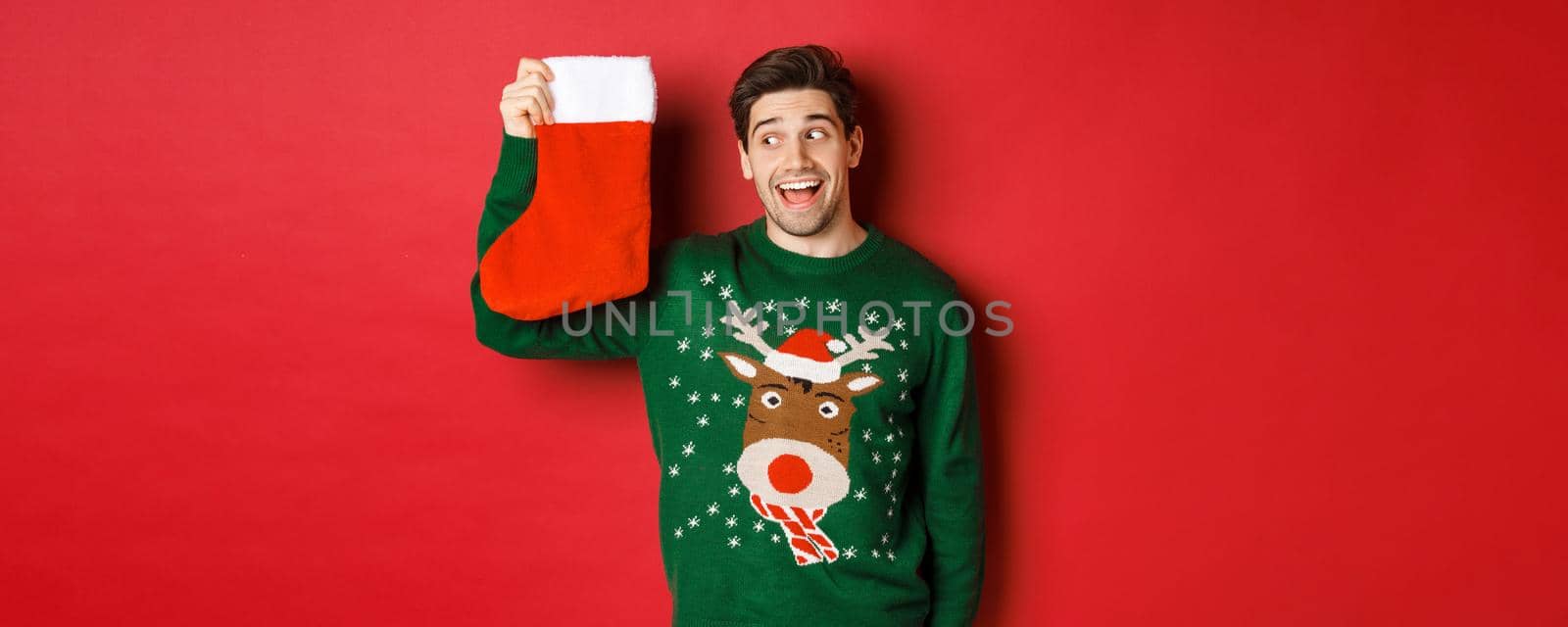 Image of surprised and amused man in green sweater, looking at christmas stocking with presents and smiling, standing over red background.