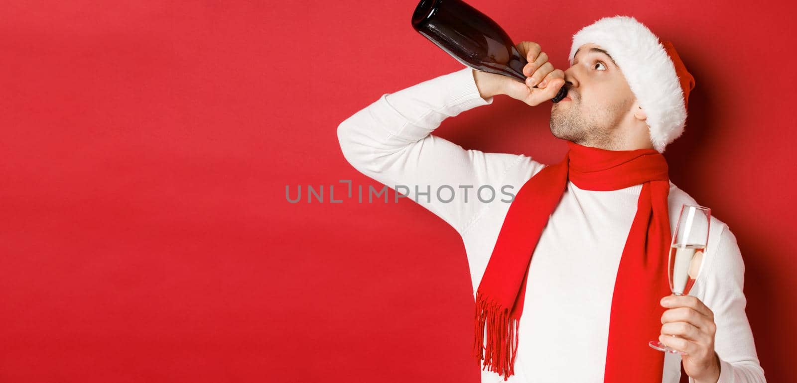 Concept of winter holidays, christmas and lifestyle. Man getting drunk on new year party, drinking champagne from bottle, wearing santa hat, standing over red background.