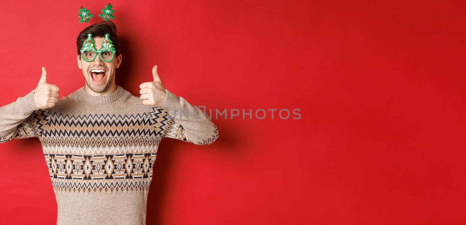 Image of excited handsome guy in party glasses and christmas sweater, showing thumbs-up, praising great new year celebration, standing over red background by Benzoix