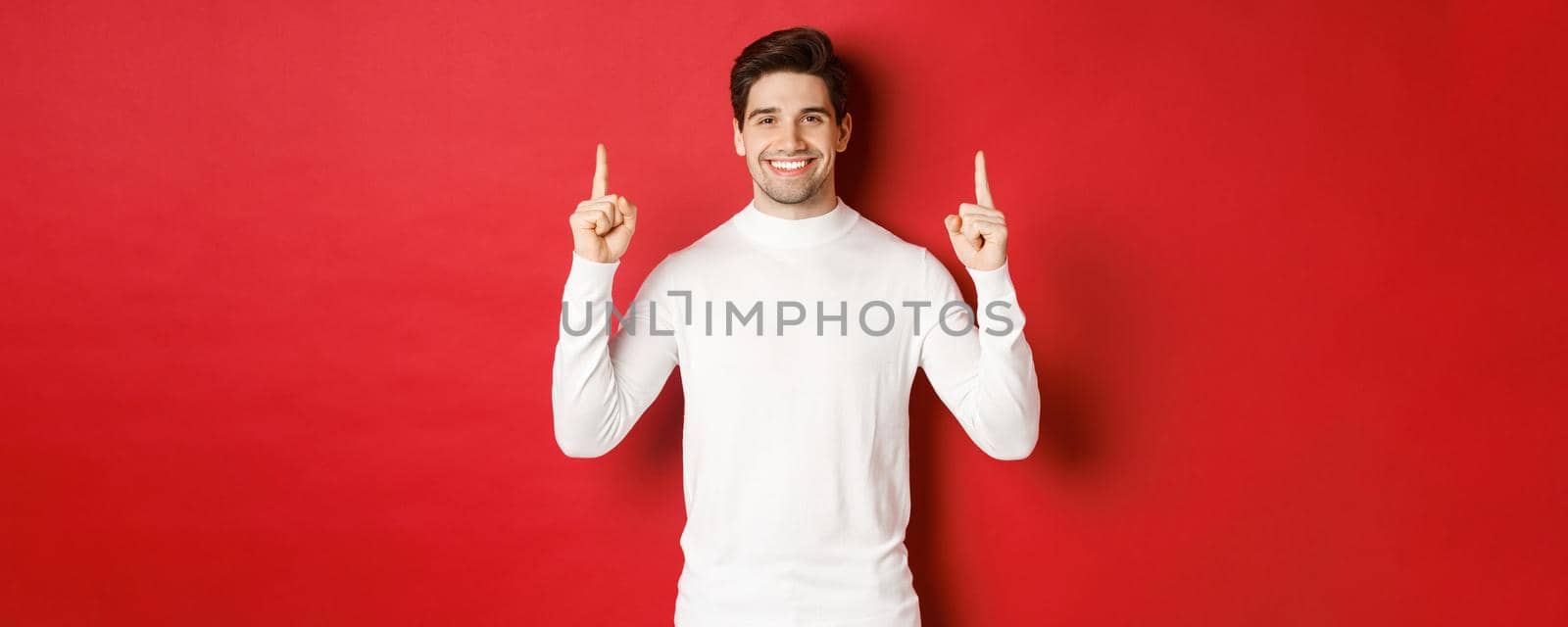 Concept of winter holidays. Good-looking young man with bristle, wearing white sweater, showing christmas advertisement on copy space, pointing fingers up and smiling, red background.