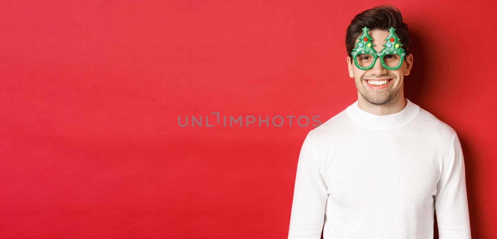 Concept of christmas, winter holidays and celebration. Close-up of handsome man in white sweater and party glasses, enjoying new year, standing over red background.