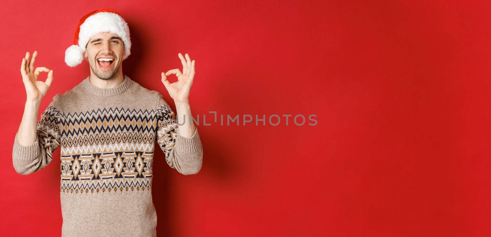 Image of joyful attractive guy in sweater and santa hat, wishing merry christmas, showing okay signs and winking at camera, celebrating new year, standing over red background.