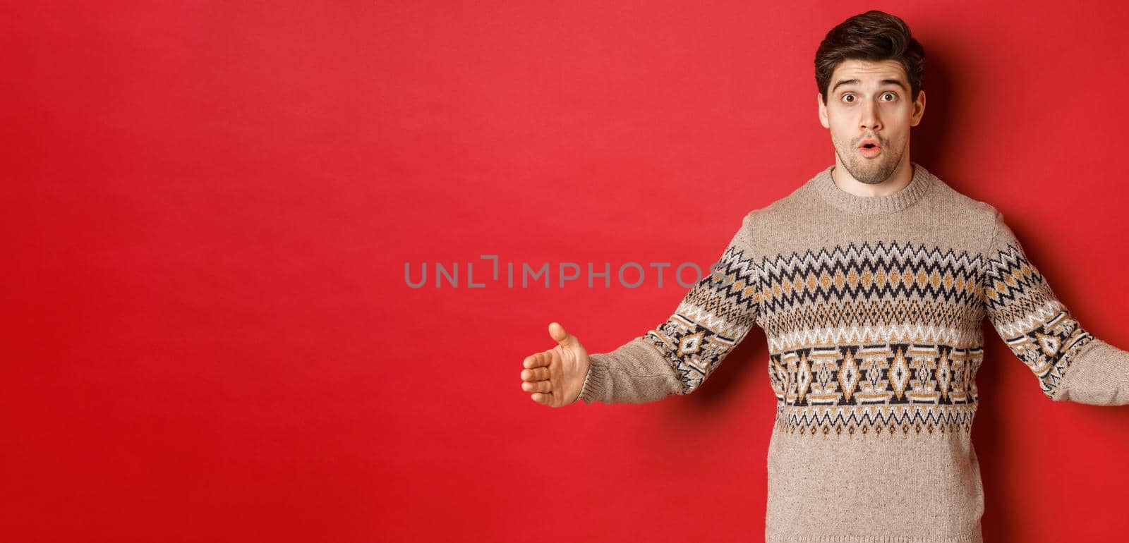 Image of amazed handsome man showing size of christmas present, spread hands sideways and shaping something large, standing over red background.
