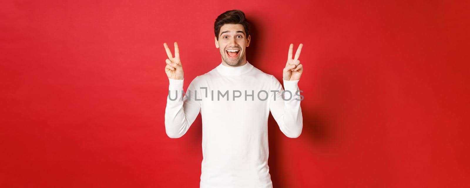Concept of winter holidays, christmas and lifestyle. Handsome funny guy in white sweater, showing peace signs and smiling happy, standing against red background.