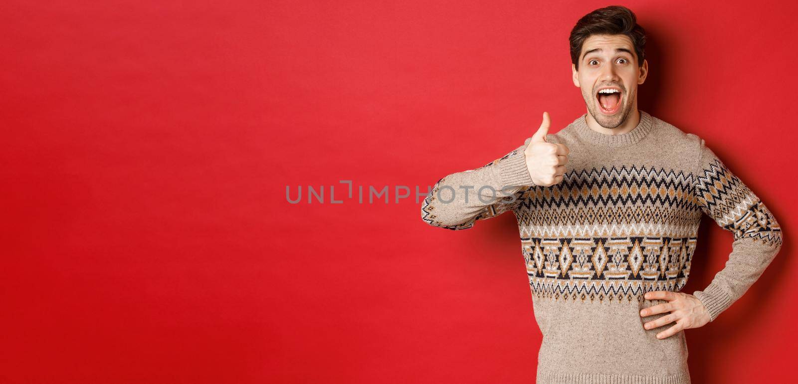 Portrait of excited, attractive man in christmas sweater, showing thumbs-up and looking amazed, recommending good thing, advertising christmas related product by Benzoix