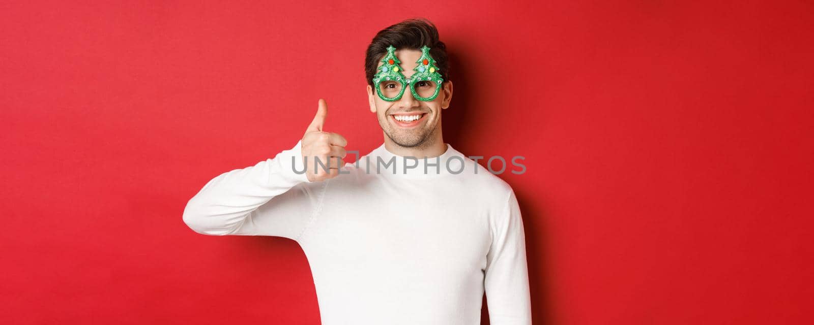 Concept of christmas, winter holidays and celebration. Close-up of handsome smiling man, wearing party glasses and white sweater, showing thumbs-up, recommending new year promo.