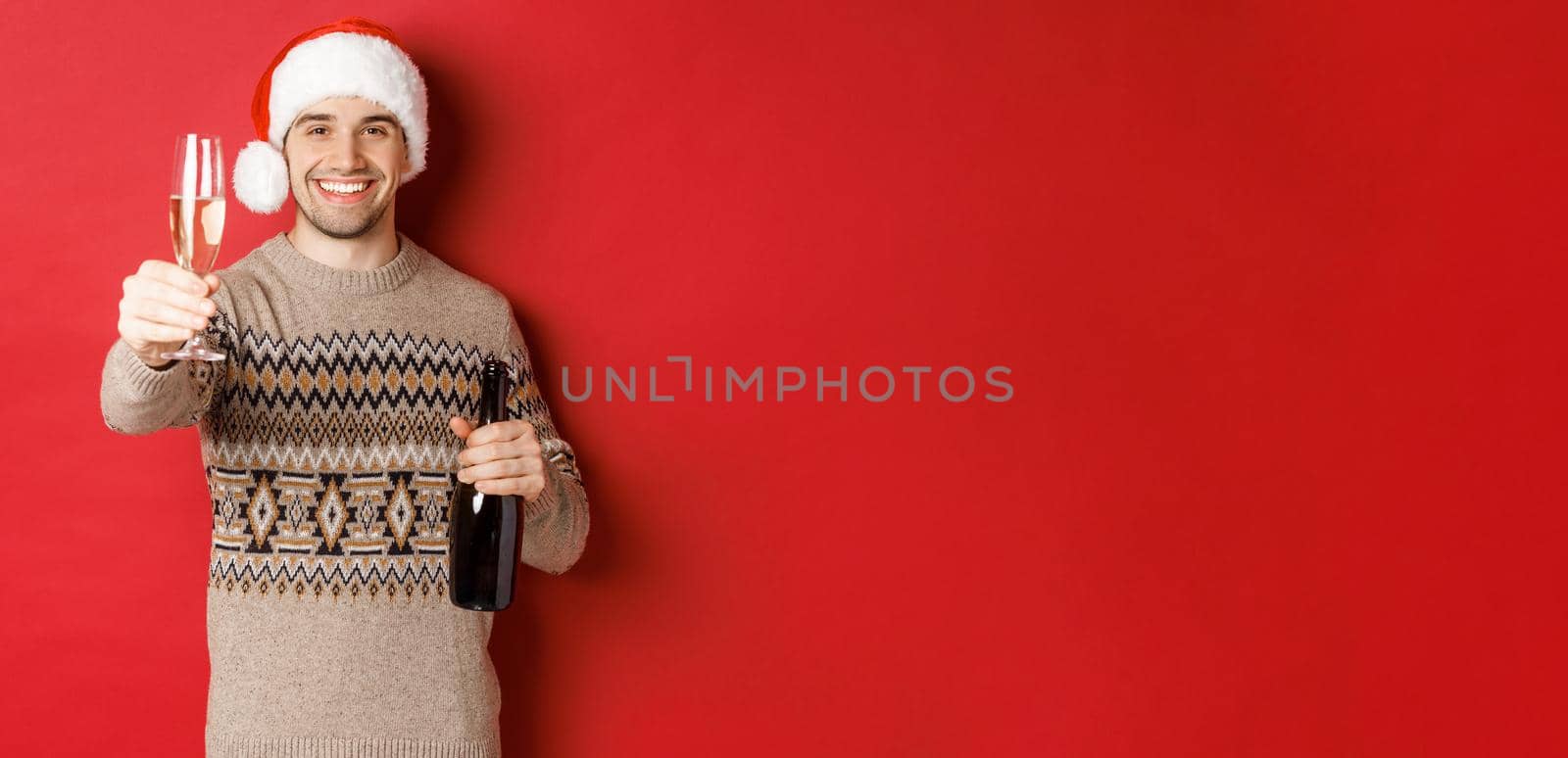 Concept of winter holidays, new year and celebration. Portrait of handsome man in santa hat and sweater, holding champagne, raising glass and saying cheers on christmas party.