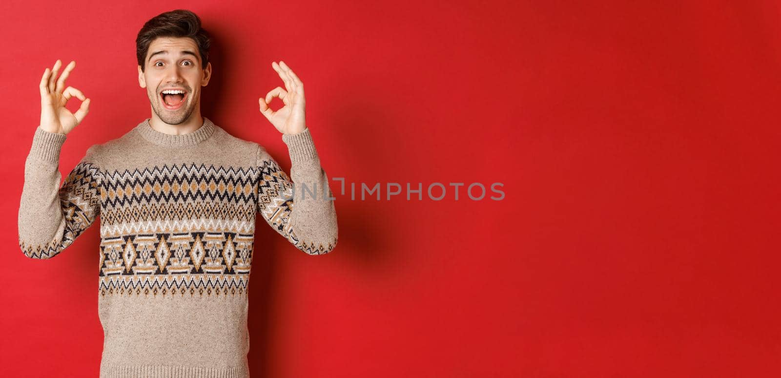 Concept of christmas celebration, winter holidays and lifestyle. Portrait of handsome man in sweater, looking amazed and showing okay signs, guarantee or recommend something good, red background by Benzoix