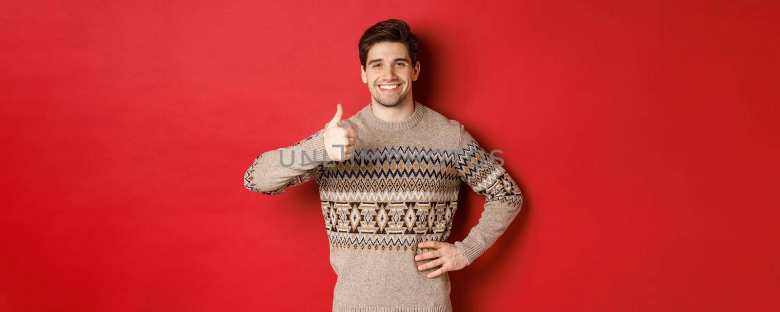 Portrait of happy handsome man in christmas sweater, smiling and looking satisfied, showing thumbs-up in approval, like new year holiday, standing over red background.