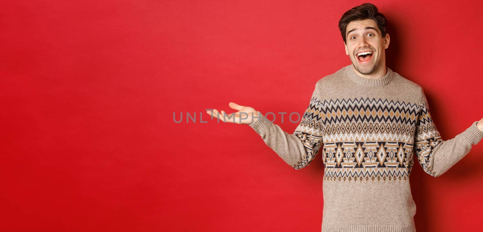 Portrait of happy and surprised, handsome caucasian guy, wearing christmas sweater, spread hands sideways and looking clueless, standing over red background by Benzoix