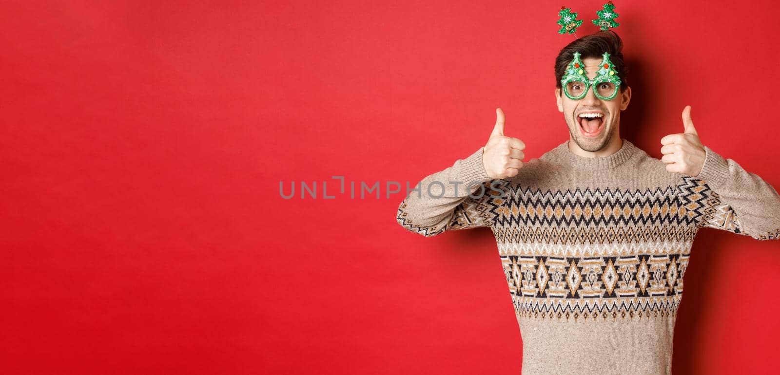 Image of excited handsome guy in party glasses and christmas sweater, showing thumbs-up, praising great new year celebration, standing over red background.