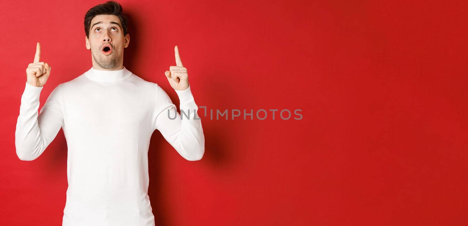 Concept of winter holidays. Portrait of handsome surprised man in white sweater, reacting to new year promo offer, looking and pointing fingers up, standing impressed against red background by Benzoix