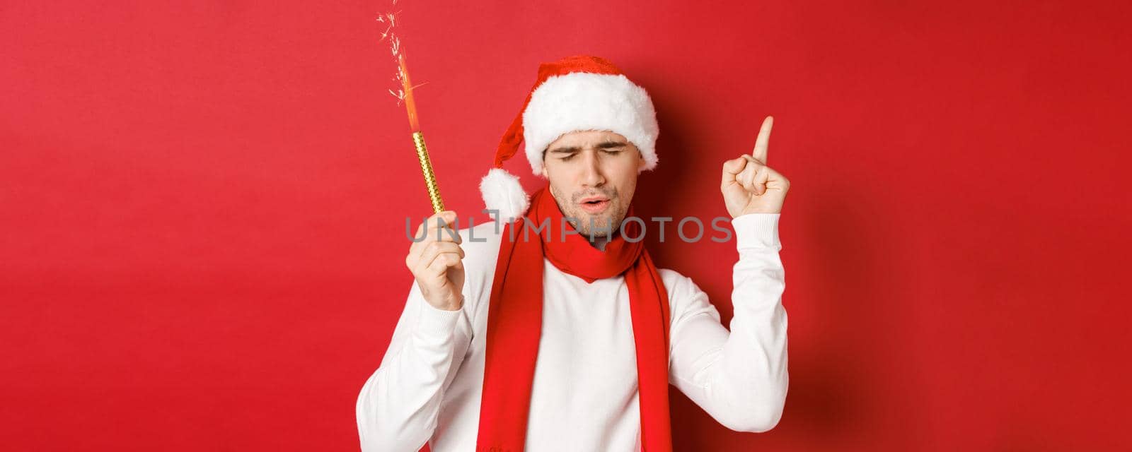 Concept of christmas, winter holidays and celebration. Attractive guy enjoying new year party, dancing with sparkler, wearing santa hat and scarf, standing over red background.