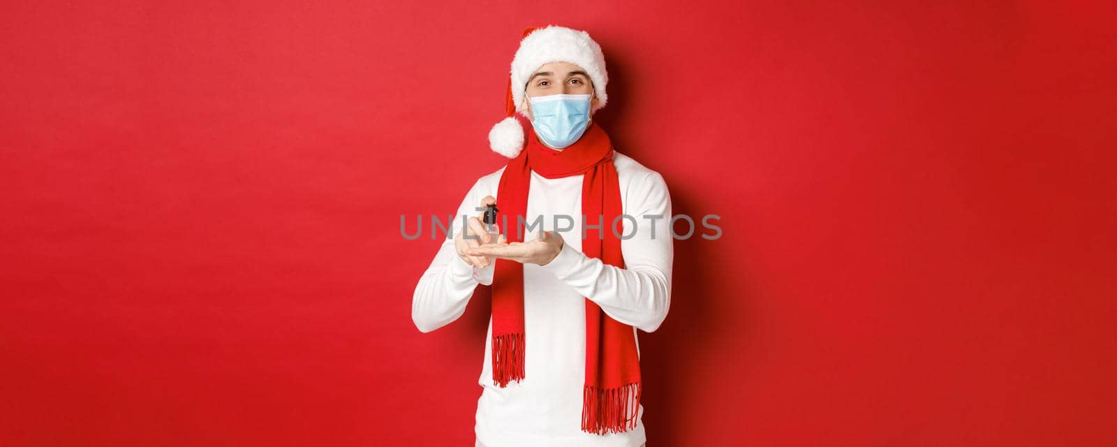 Concept of covid-19, christmas and holidays during pandemic. Happy young man in santa hat and medical mask, sanitize hands with antiseptic and smiling, standing over red background by Benzoix