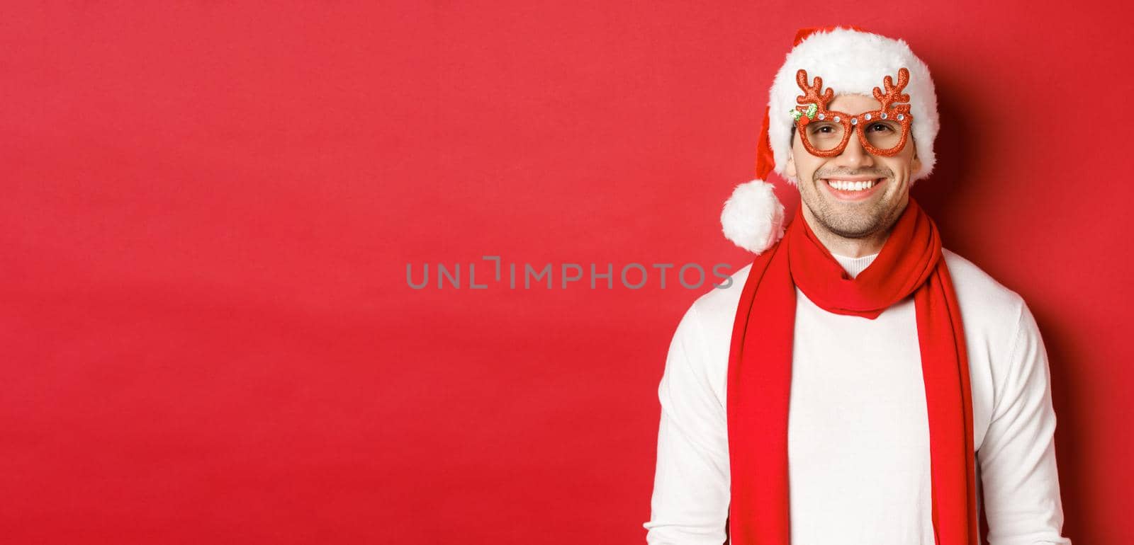Concept of christmas, winter holidays and celebration. Cheerful handsome guy in new year party glasses and smiling, standing over red background by Benzoix