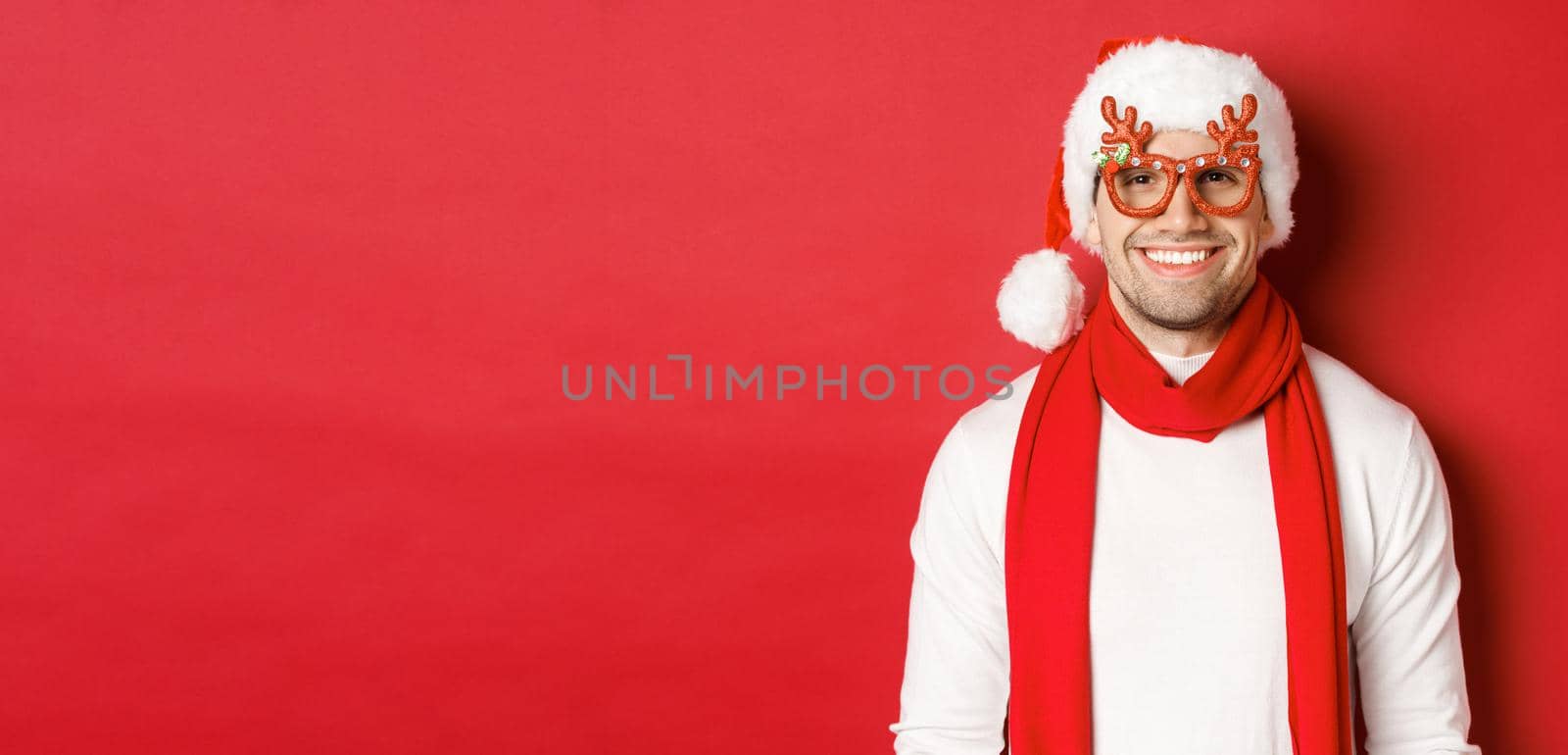 Concept of christmas, winter holidays and celebration. Cheerful handsome guy in new year party glasses and smiling, standing over red background.