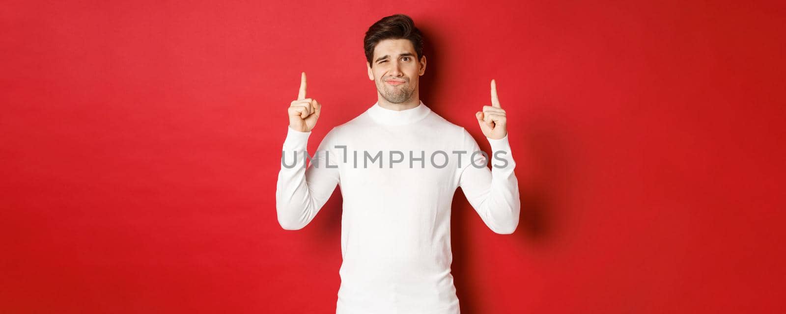 Concept of winter holidays. Disappointed handsome man do not recommend promo, pointing fingers up and grimacing displeased, standing over red background in white sweater by Benzoix