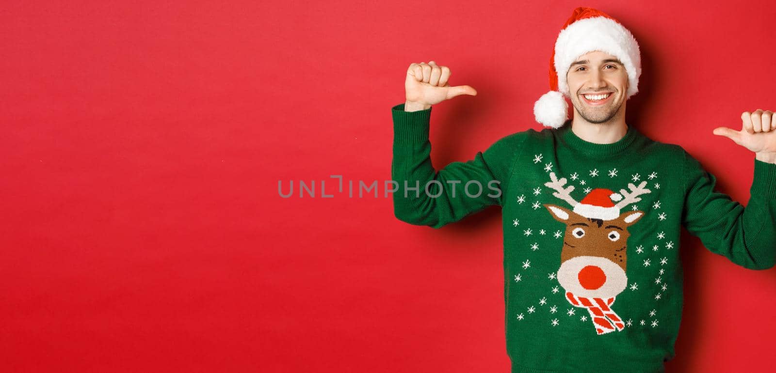 Concept of winter holidays, christmas and lifestyle. Happy attractive guy in santa hat and sweater, pointing at himself with pleased face, standing over red background by Benzoix