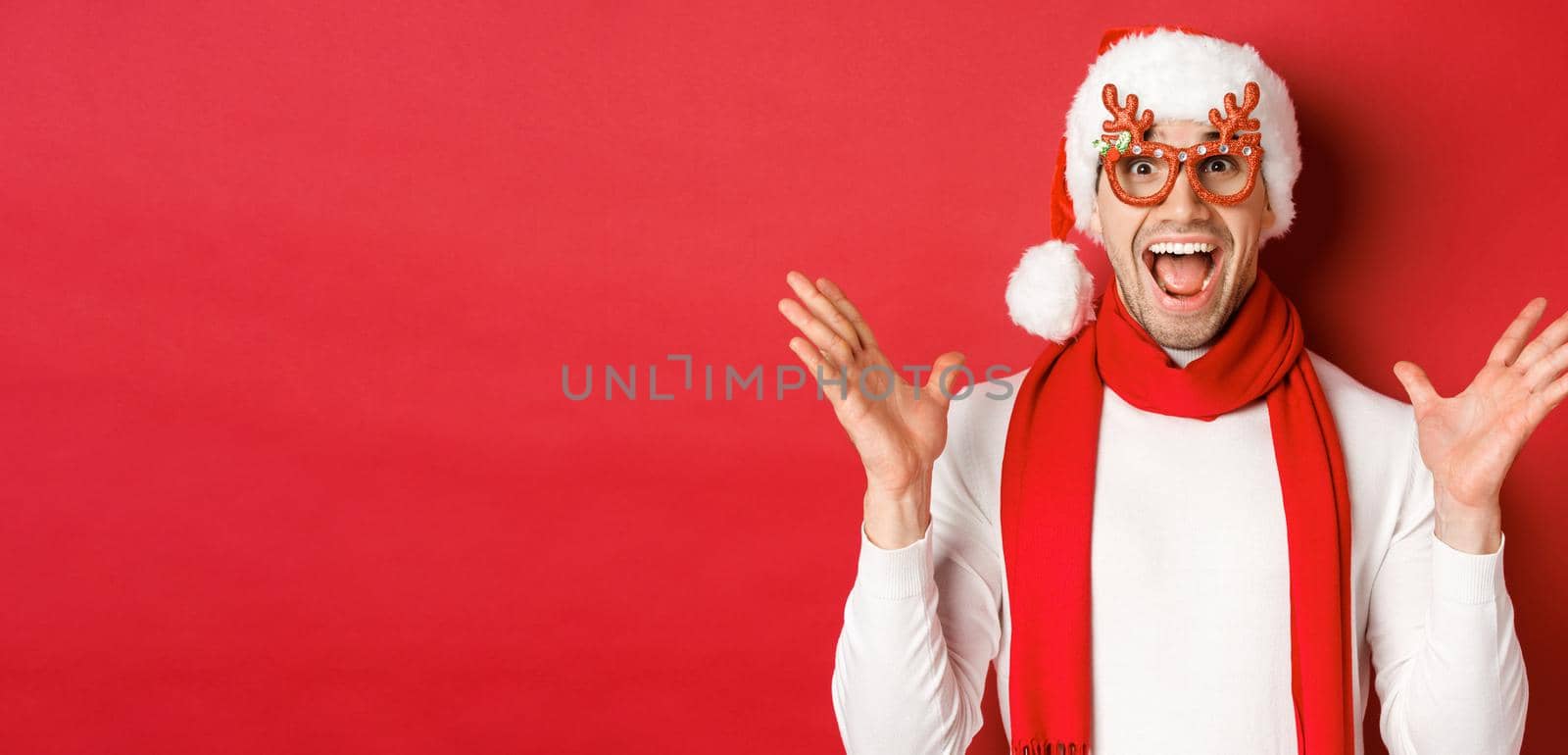 Concept of christmas, winter holidays and celebration. Image of surprised and happy man looking amazed, wearing party glasses and enjoying new year, standing over red background by Benzoix