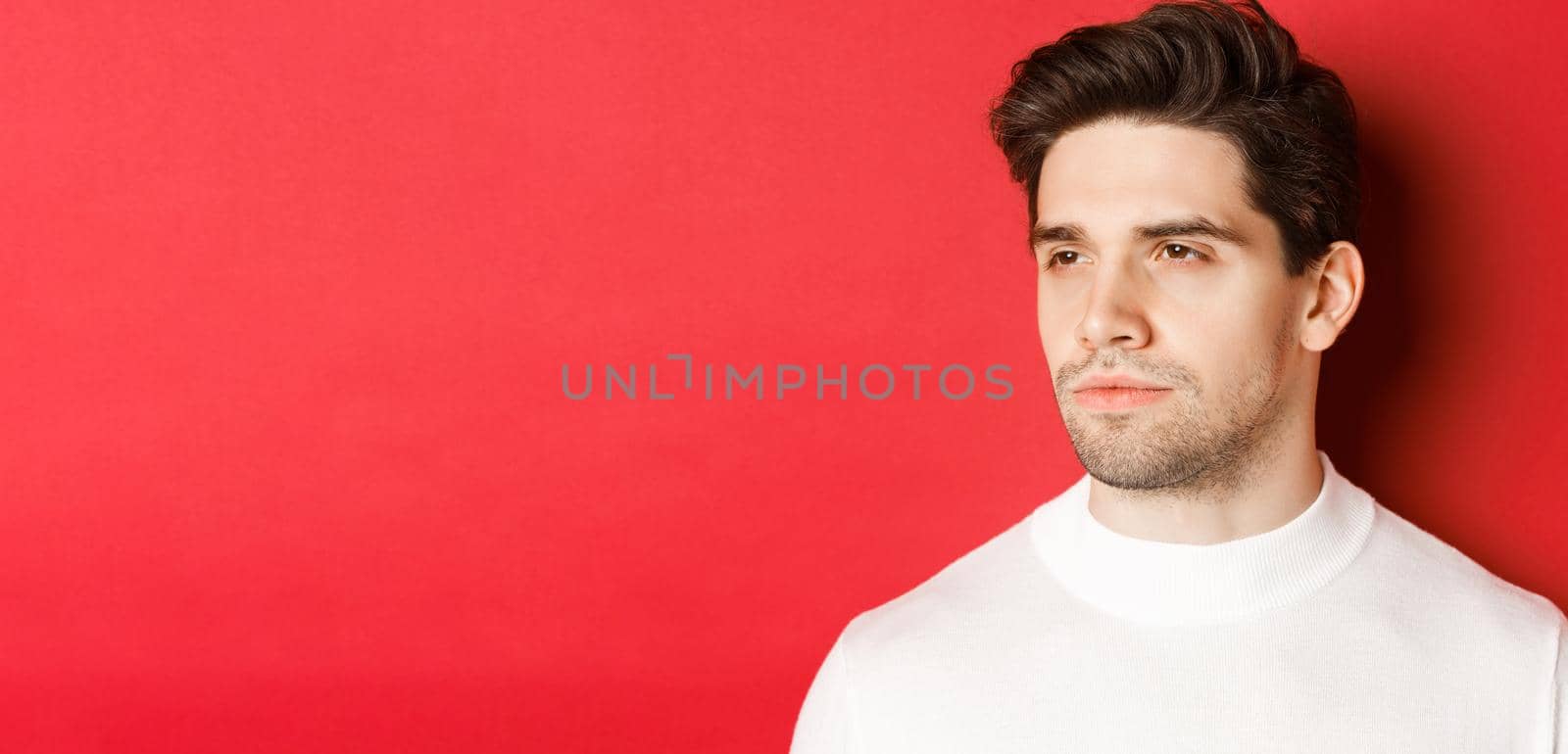Close-up of serious and thoughtful man in white sweater, looking left, standing against red background by Benzoix