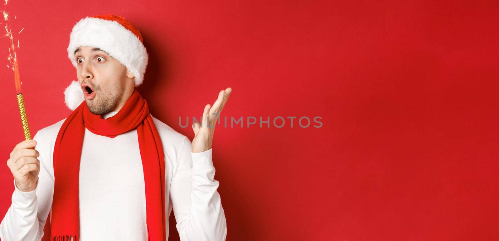 Concept of christmas, winter holidays and celebration. Close-up of amazed handsome man in santa hat and scarf, looking at sparkler with excitement, standing over red background by Benzoix