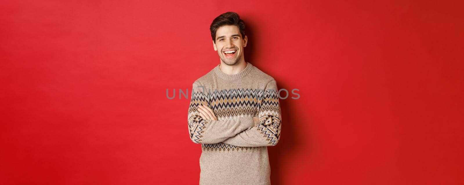 Portrait of cheerful, attractive man in christmas sweater, laughing and smiling, celebrating new year and winter holidays, standing over red background by Benzoix