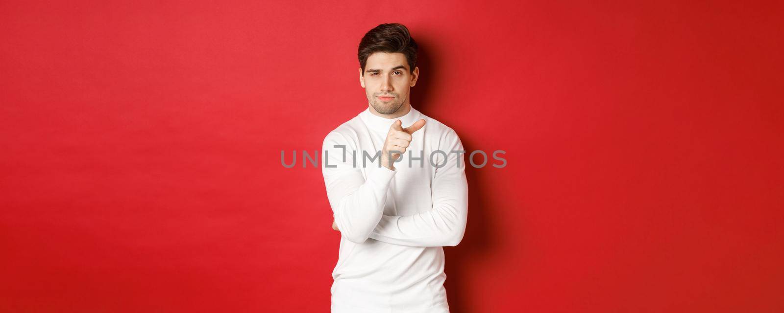 Portrait of thoughtful handsome man in white sweater, pointing at camera and making choice, standing against red background.