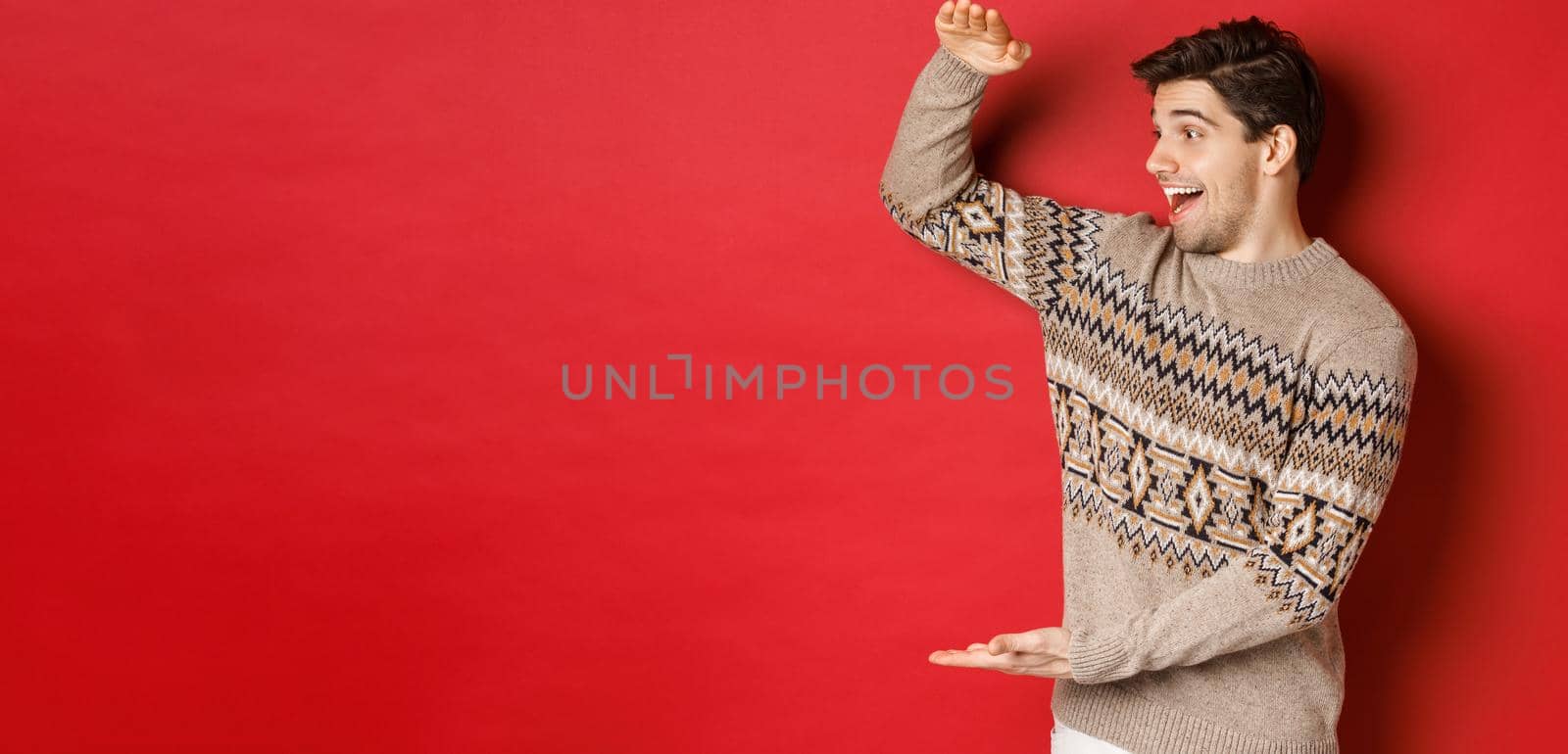 Portrait of happy young man in christmas sweater, showing large present, smiling and looking amazed at cool gift, standing over red background.