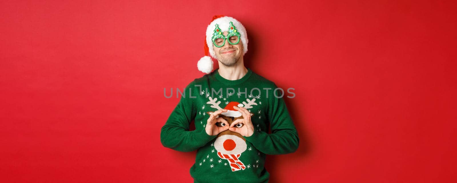 Image of happy smiling man in party glasses and santa hat, fooling around with funny christmas sweater, celebrating winter holidays, standing over red background.