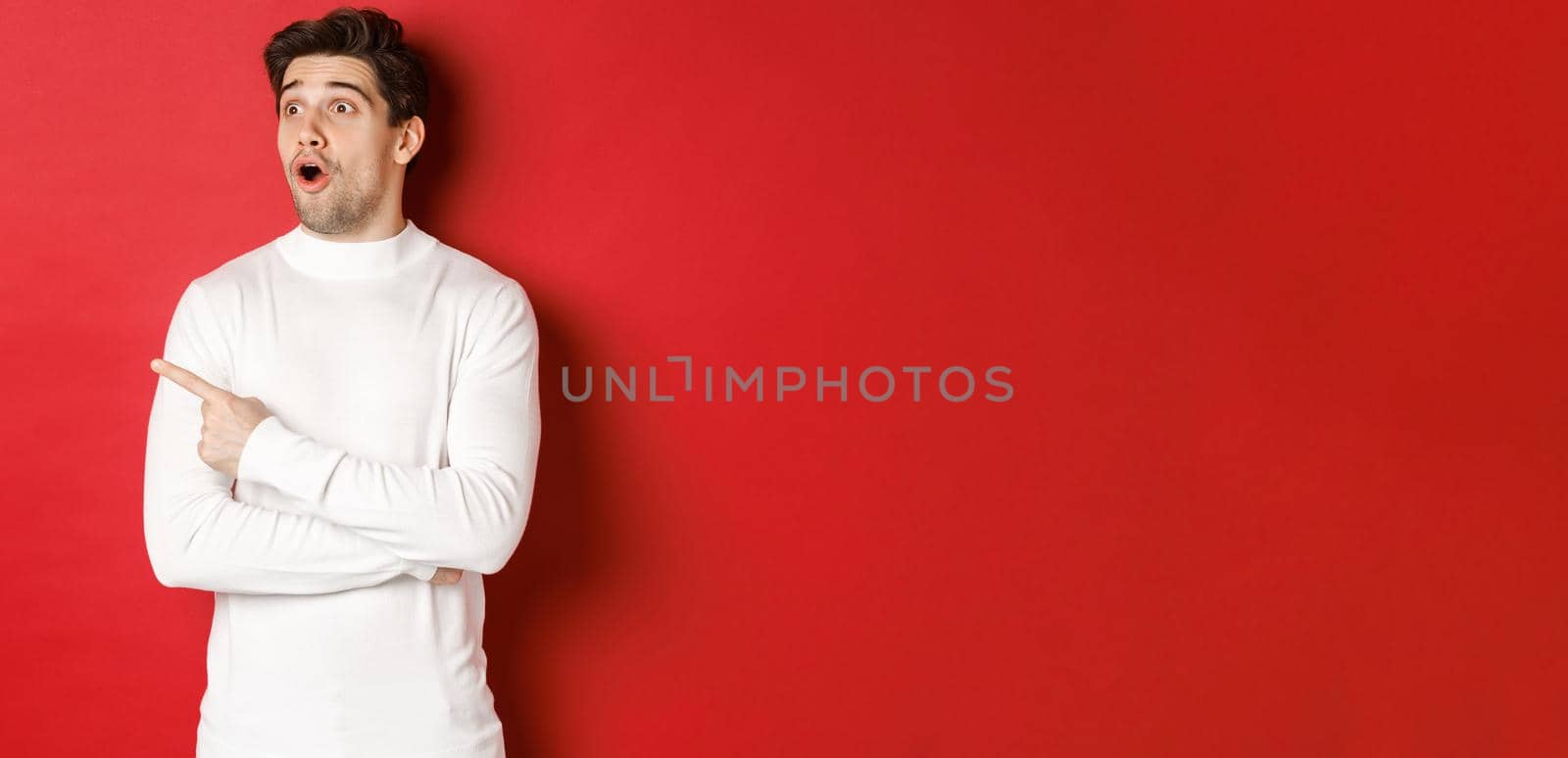Concept of winter holidays. Portrait of impressed handsome man in white sweater, pointing and looking left with dropped jaw and amazed expression, standing against red background.