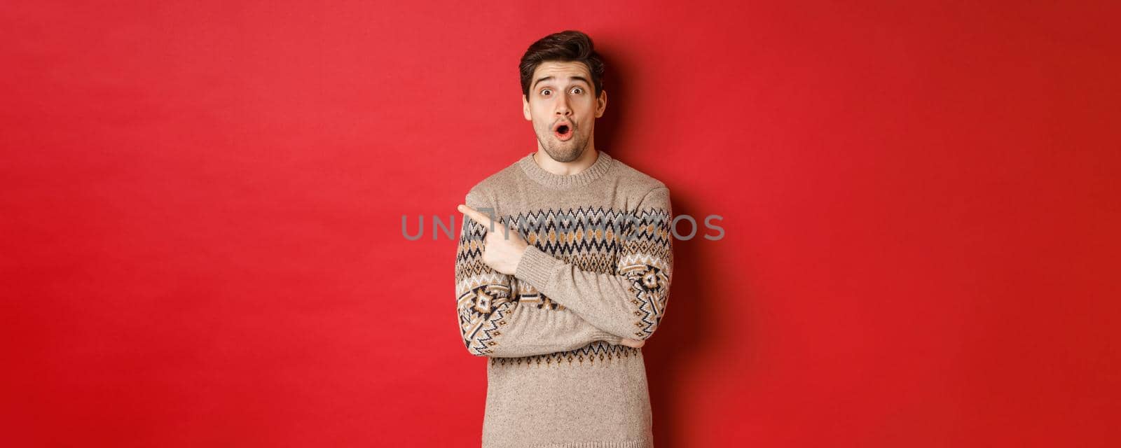 Image of impressed handsome guy, showing awesome christmas promo offer, pointing finger at upper left corner and looking amazed, standing in xmas sweater over red background.