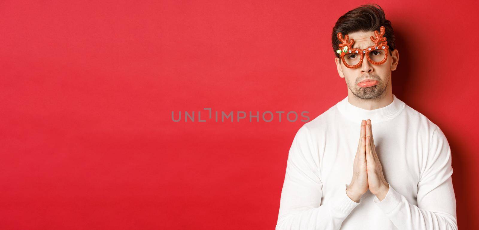 Image of handsome man in christmas party glasses, begging for help or apologizing, need a favour, standing over red background by Benzoix